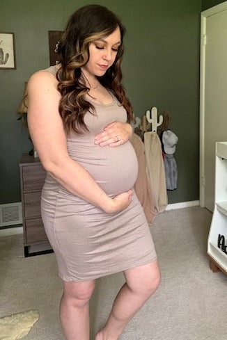 woman wearing a beige fitted maternity dress and cradling her baby bump while standing in a well-lit room