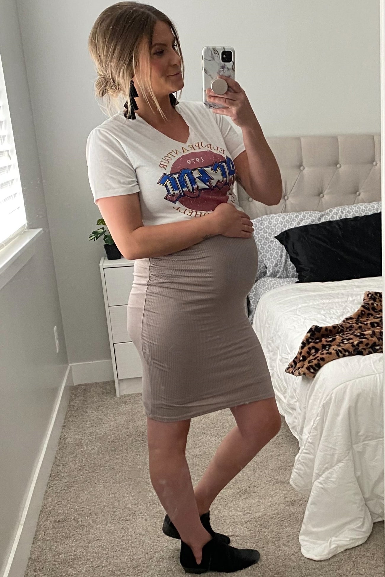 woman wearing a graphic tee and a beige fitted maternity skirt, holding her baby bump in a bright bedroom