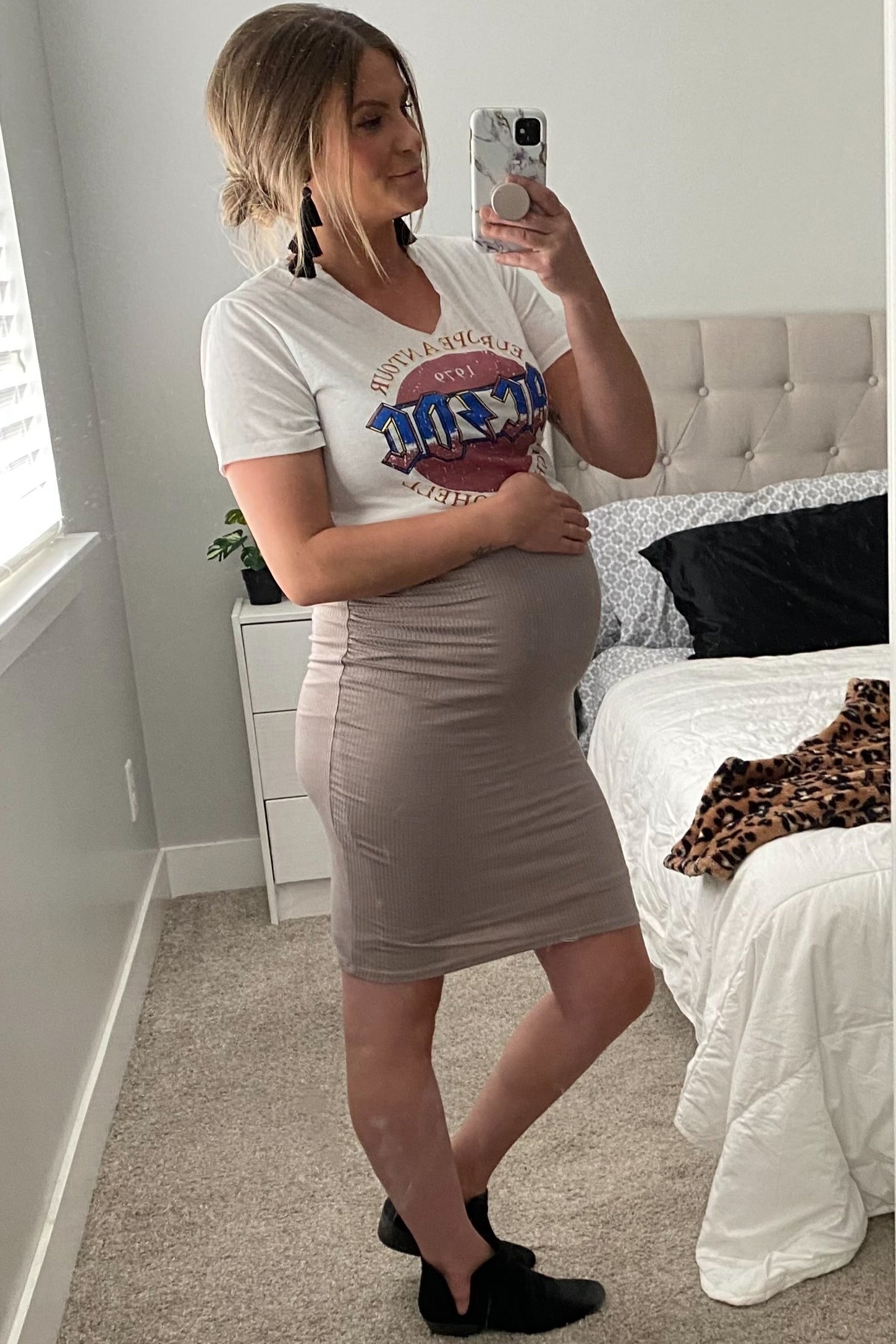 woman wearing a graphic tee and a beige fitted maternity skirt, holding her baby bump in a bright bedroom