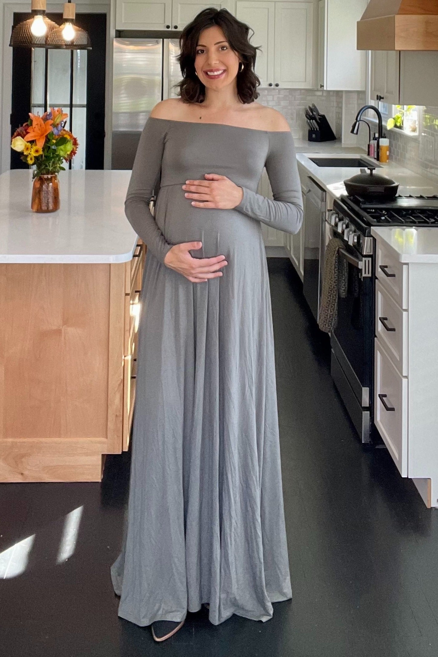 woman wearing a long sleeve gray maternity dress and holding her baby bump in a kitchen setting