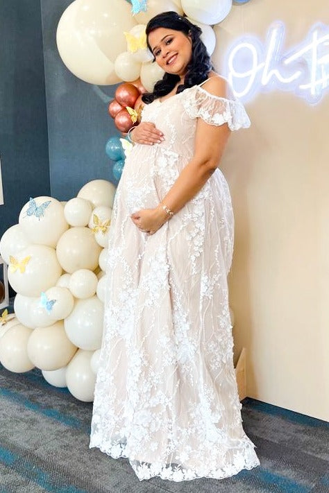 smiling pregnant woman in white lace maternity dress posing at baby shower with balloon backdrop