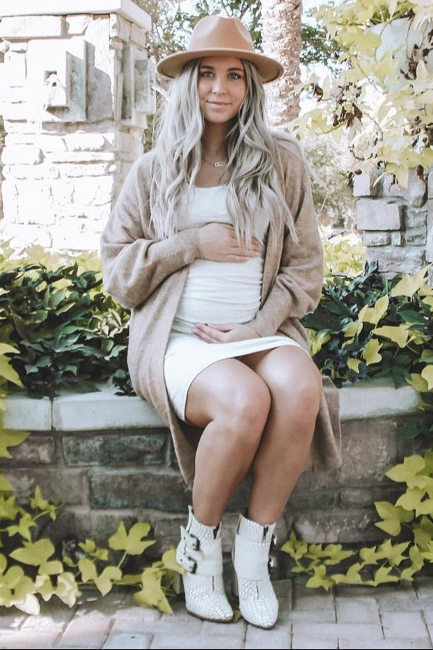 woman wearing a cream maternity dress and a tan cardigan, sitting outdoors and gently holding her baby bump