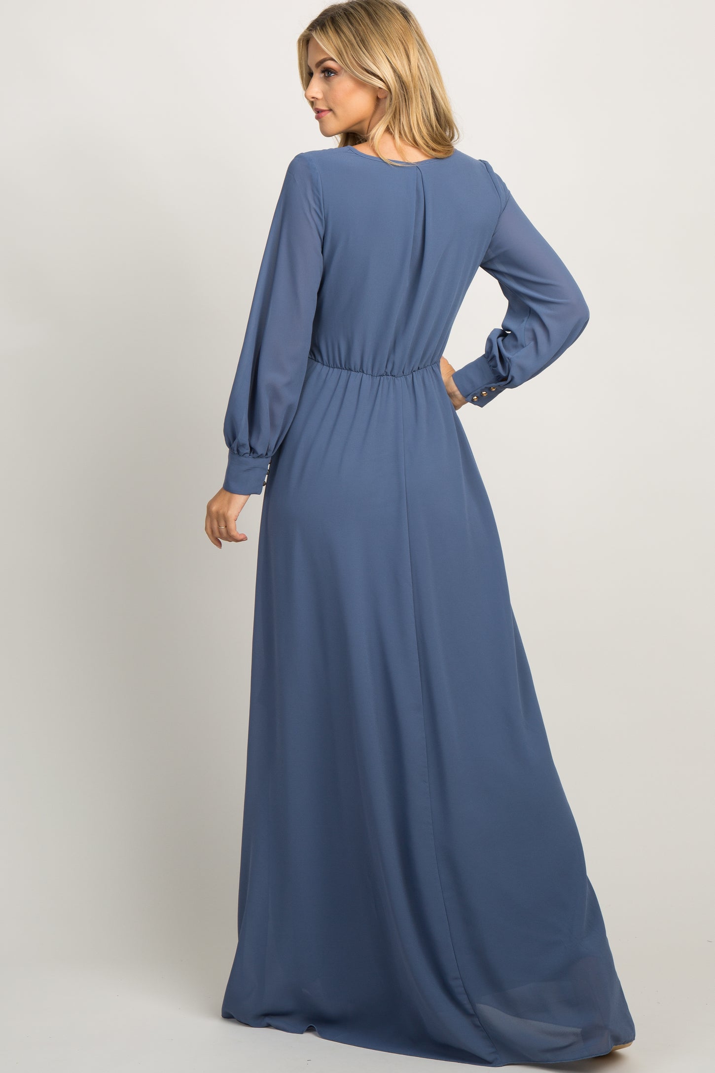 Woman wearing a long sleeve slate blue chiffon maxi dress with a pleated waist and button cuffs