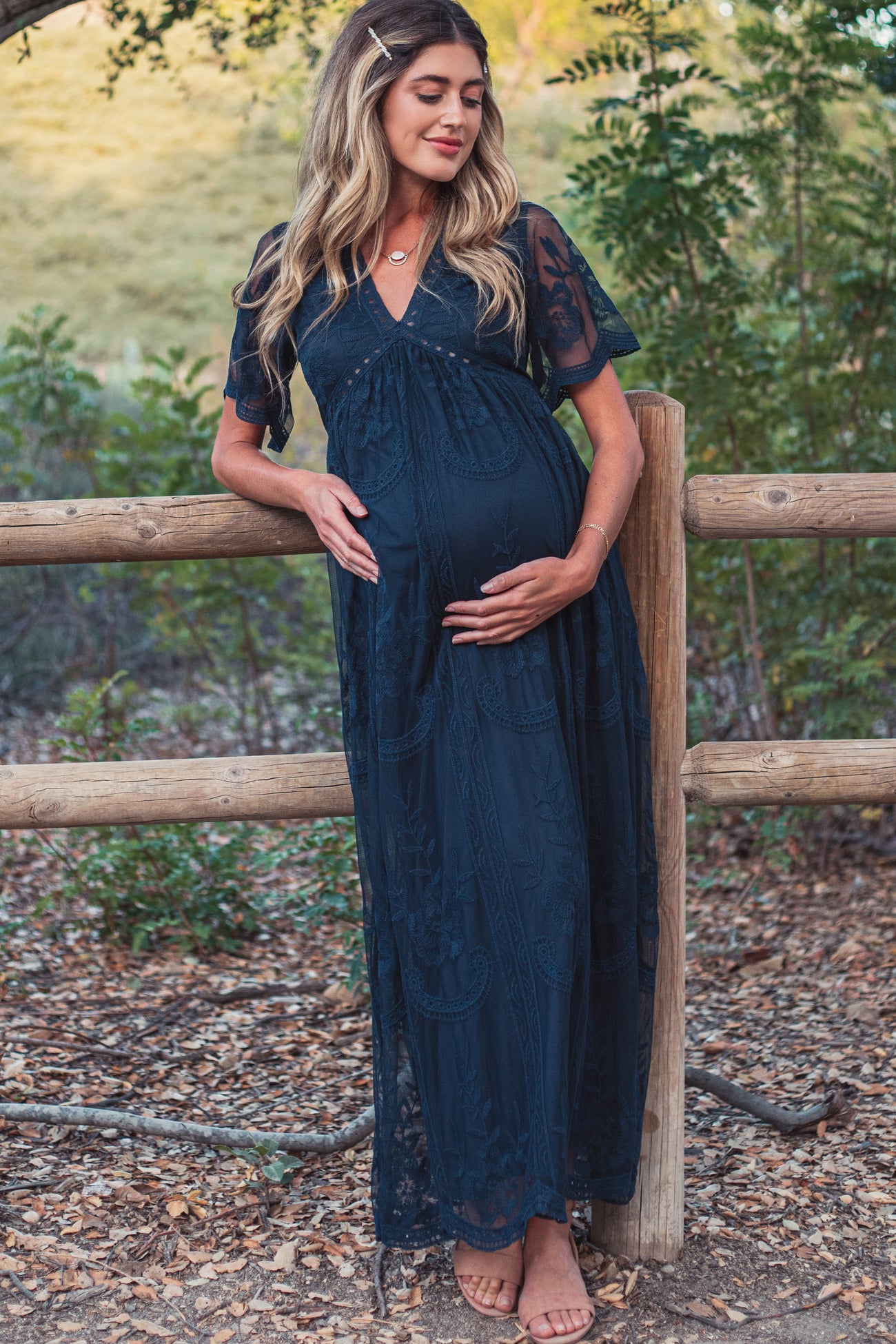 woman wearing a dark blue lace maternity maxi dress and holding her baby bump while standing by a wooden fence