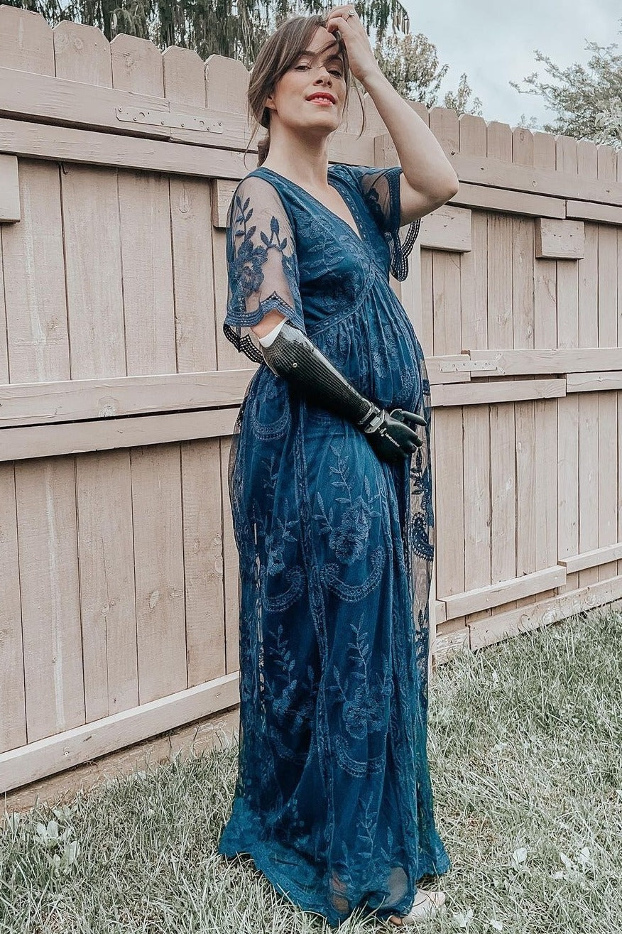 woman wearing a navy blue maternity dress with sheer sleeves, posing with her baby bump visible against a wooden fence