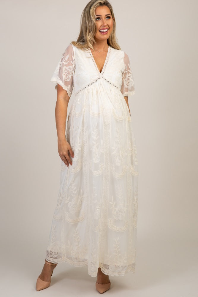 woman wearing a cream lace maternity dress with flutter sleeves and a baby bump, standing outdoors in a garden setting