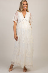 woman wearing a cream lace maternity dress with flutter sleeves and a baby bump, standing outdoors in a garden setting