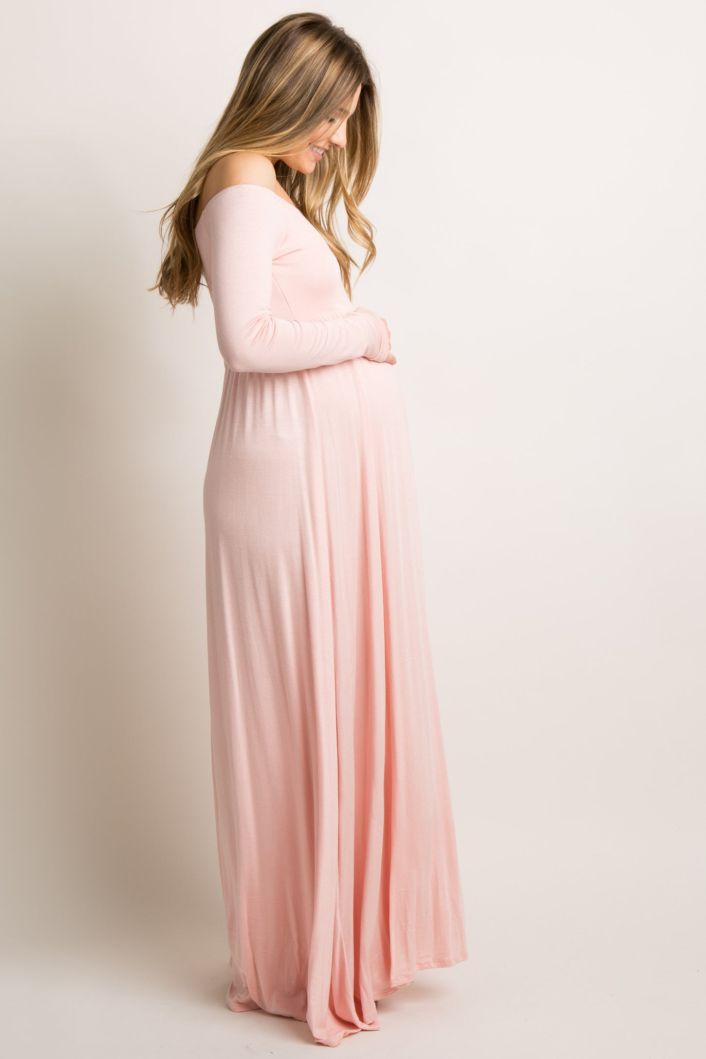 woman wearing a light pink off-the-shoulder maternity maxi dress and gently holding her baby bump