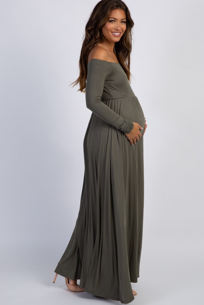 woman wearing a green off-shoulder maternity dress with a flowing skirt, cradling her baby bump while sitting in a chair
