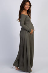 woman wearing a green off-shoulder maternity dress with a flowing skirt, cradling her baby bump while sitting in a chair