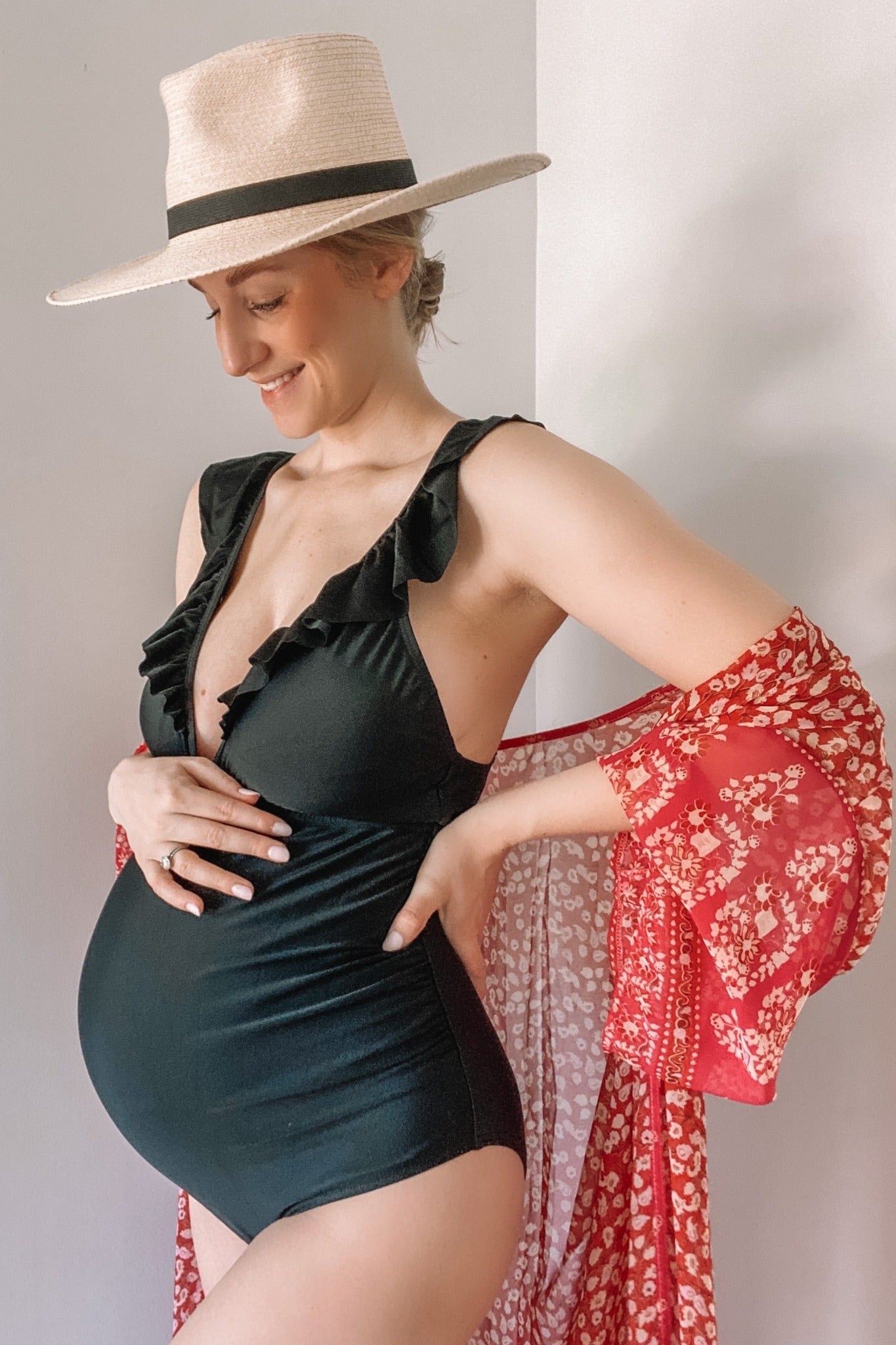woman wearing a black maternity swimsuit with ruffle details and a red sheer cover-up, holding her baby bump