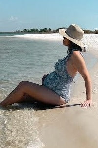 woman wearing a blue floral maternity swimsuit sitting by the water with a visible baby bump and a sun hat