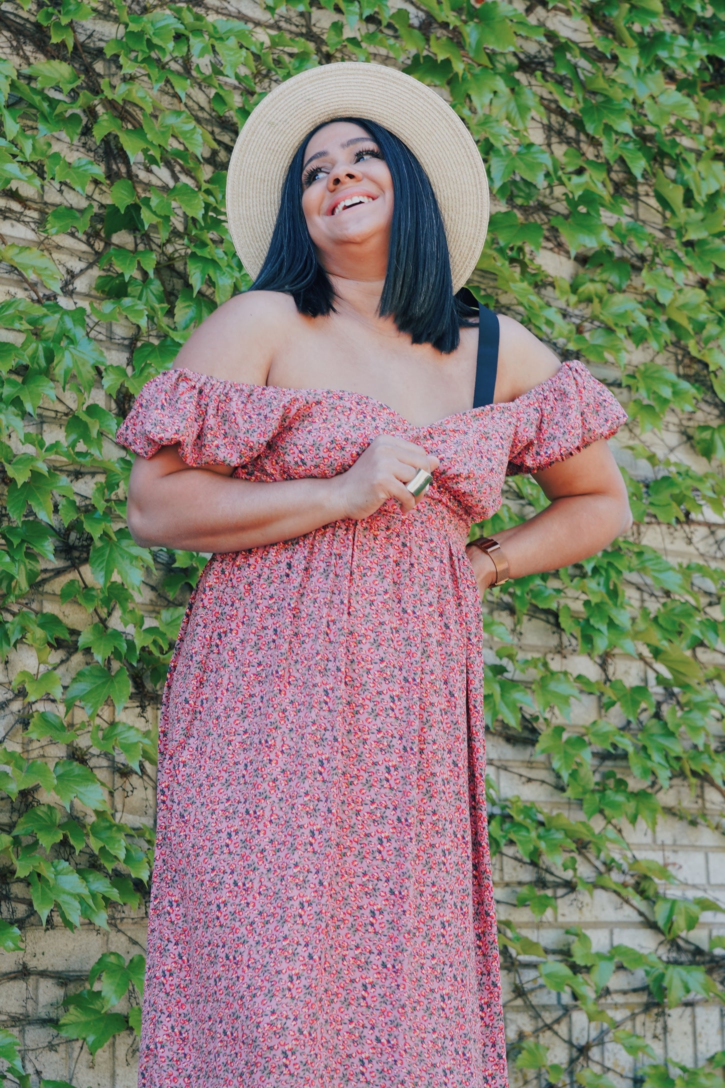 woman wearing a pink floral off-the-shoulder maxi dress and straw hat, smiling against a green leafy background