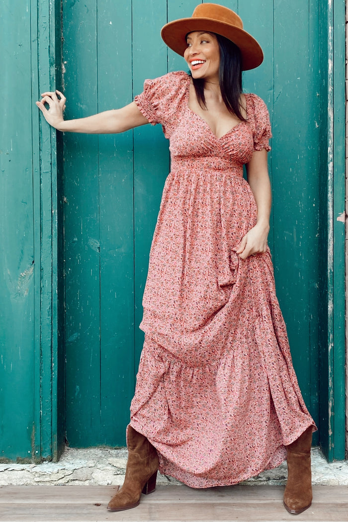 woman wearing a pink floral maxi dress with puff sleeves and a deep v-neck, accessorized with a brown wide-brimmed hat and tan boots