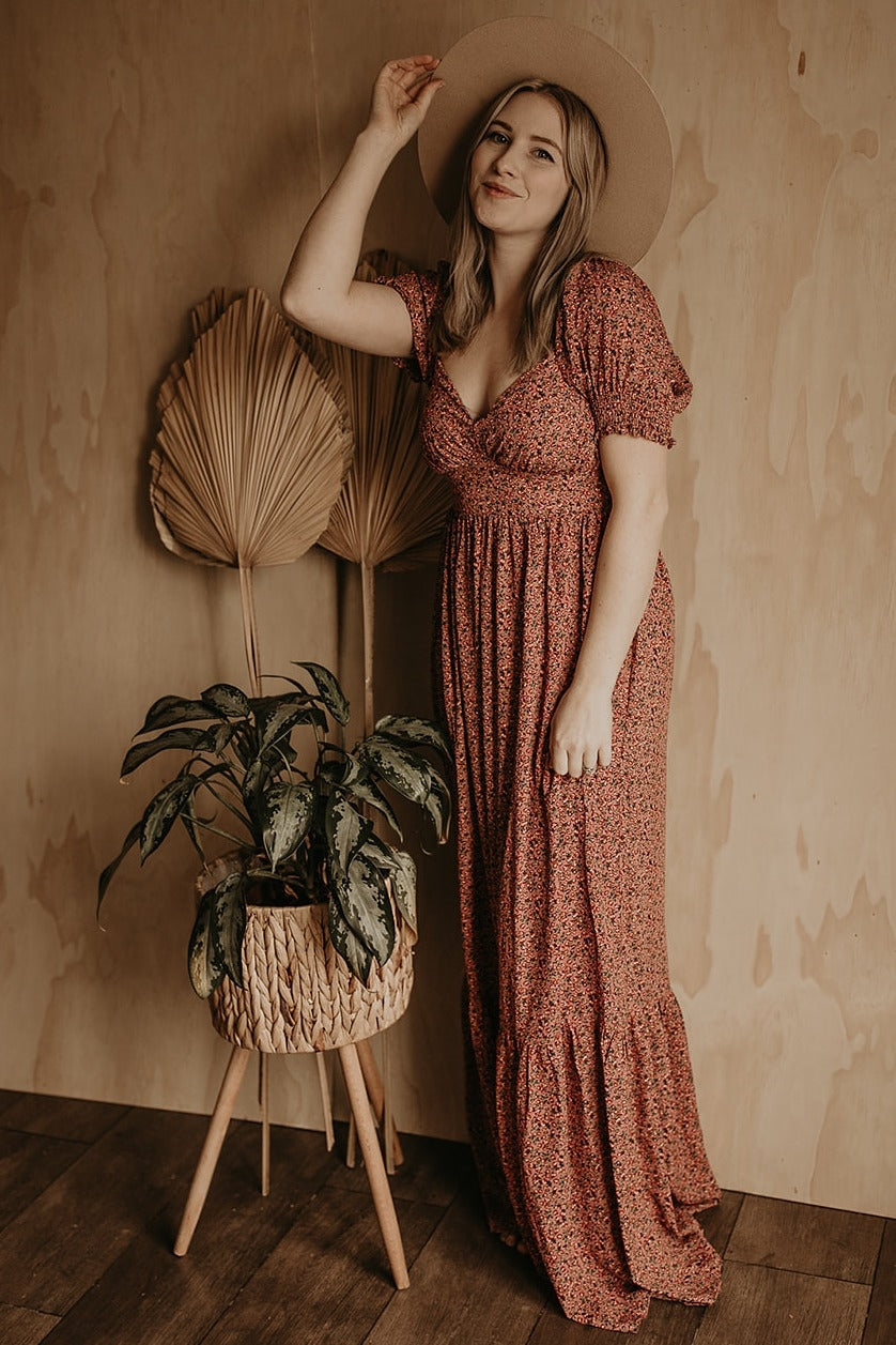 woman in floral maxi dress and wide brim hat standing by a potted plant against a wood wall