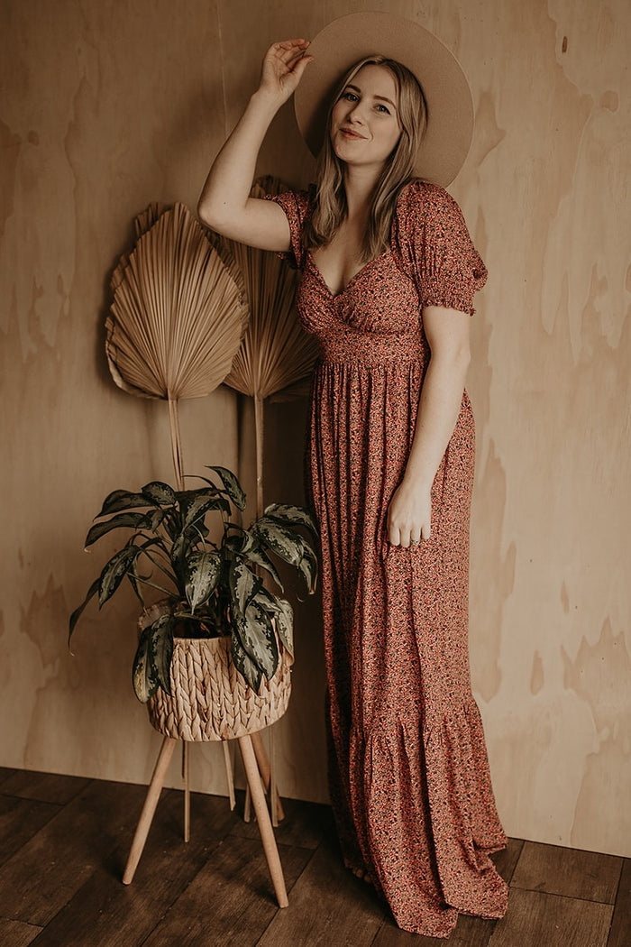 woman in floral maxi dress and wide brim hat standing by a potted plant against a wood wall