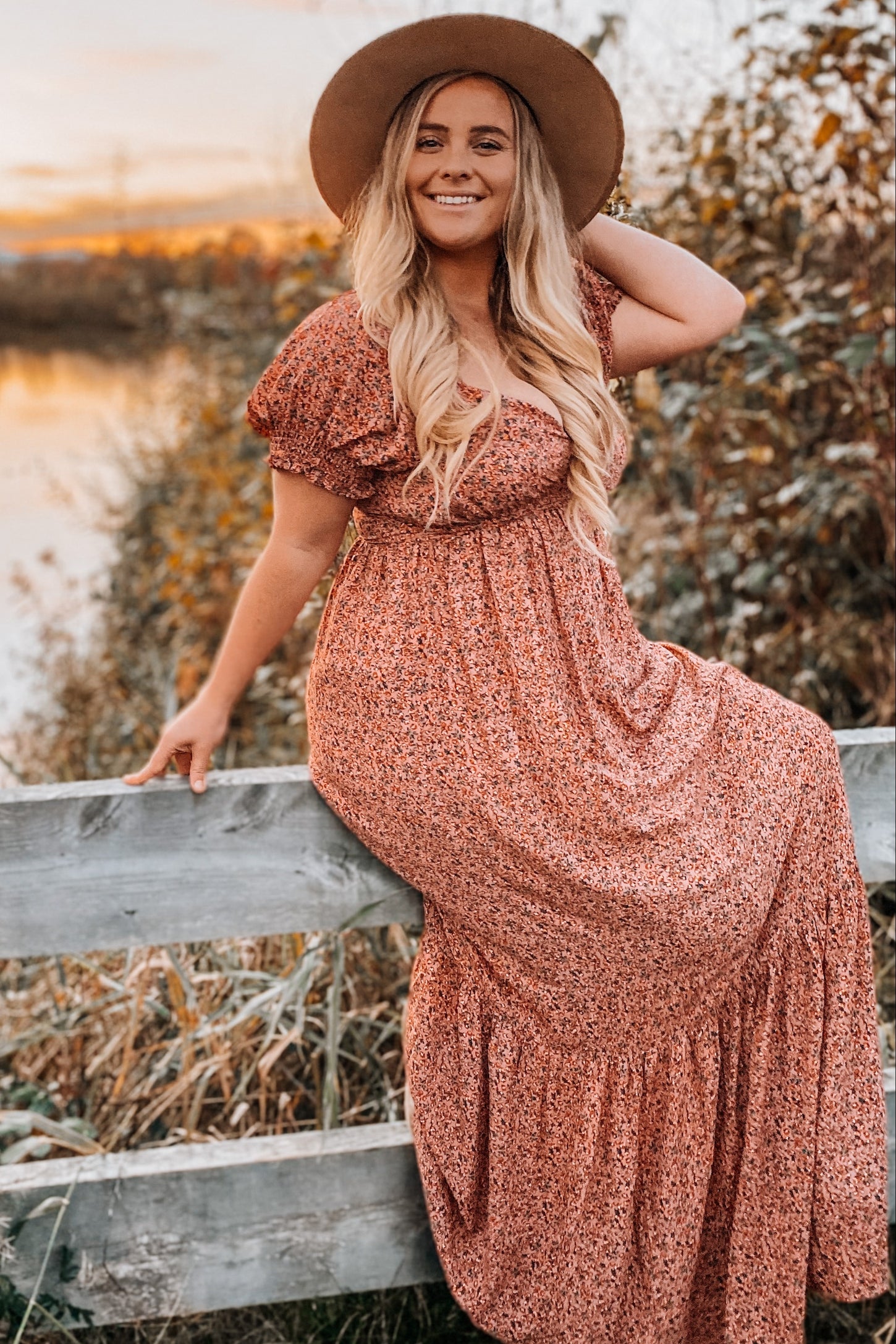 woman wearing a floral print maxi dress with short ruffled sleeves and a wide-brimmed hat, posing on a fence in a natural setting