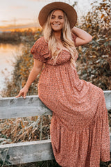 woman wearing a floral print maxi dress with short ruffled sleeves and a wide-brimmed hat, posing on a fence in a natural setting