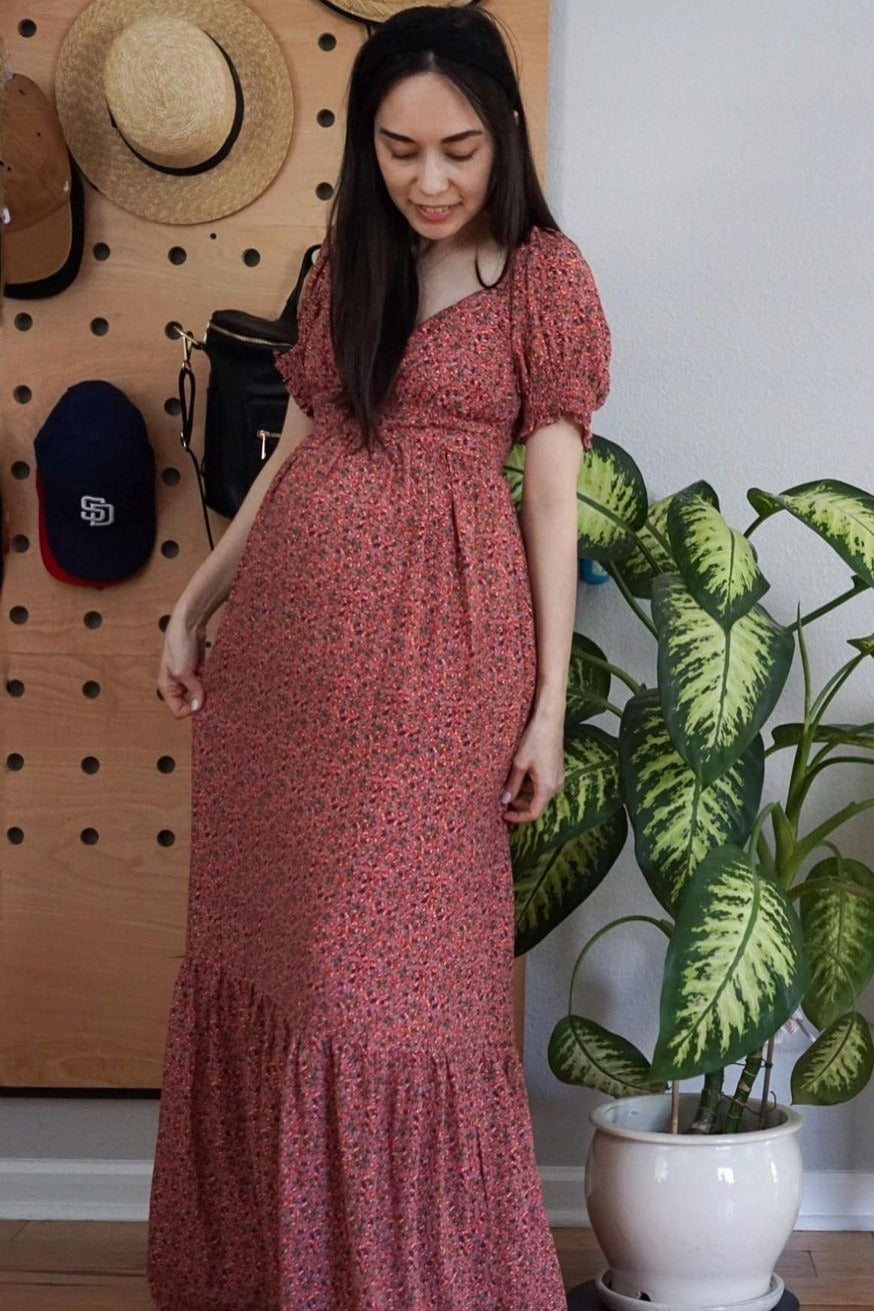 woman wearing a floral maternity maxi dress with puff sleeves and a visible baby bump, standing next to a plant
