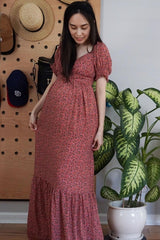 woman wearing a floral maternity maxi dress with puff sleeves and a visible baby bump, standing next to a plant