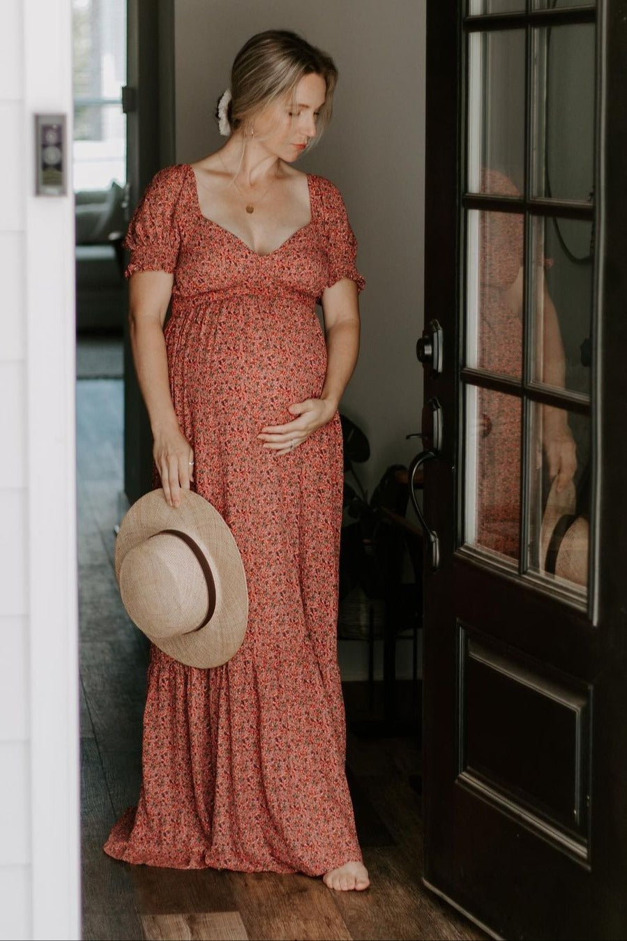 woman wearing a red floral maternity dress with puff sleeves, holding a straw hat and cradling her baby bump