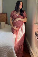 woman wearing a floral pink maternity dress cradling her baby bump while standing by a window
