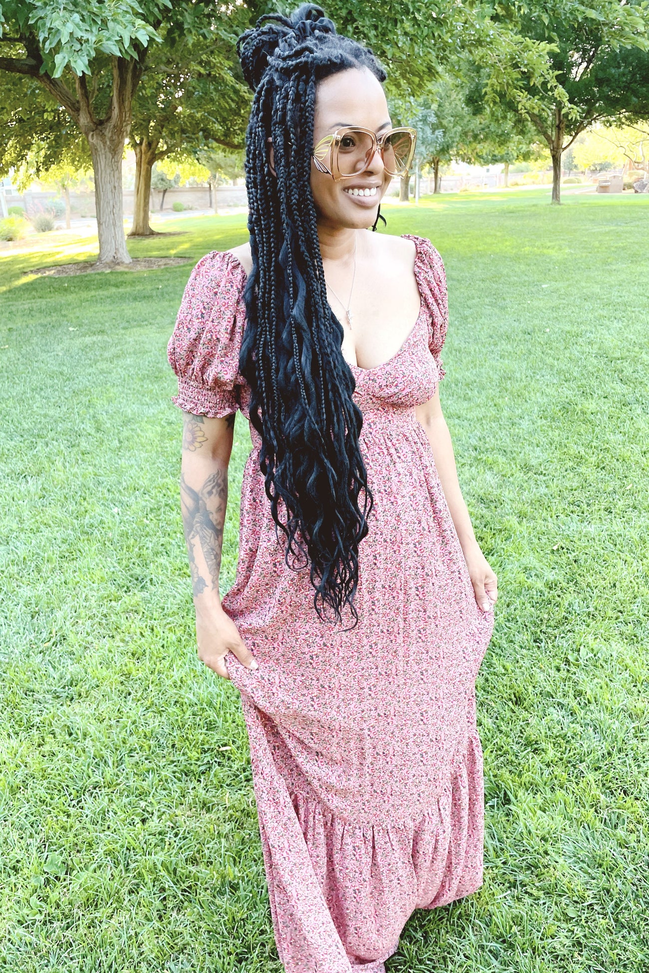 woman with long black braids wearing a pink floral maxi dress and oversized sunglasses in a park