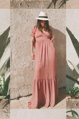 Woman in a pink floral maxi dress and white fedora hat leaning against a stone wall next to agave plants