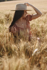 woman wearing a floral brown dress and a wide-brimmed hat, posing in a field of tall wheat grass