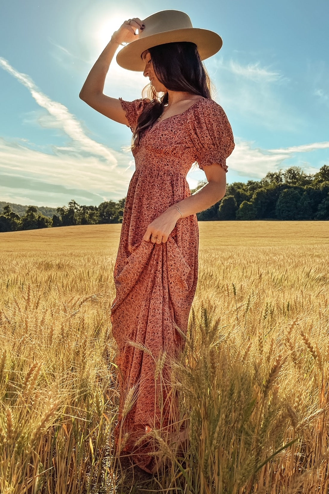 woman wearing a floral maxi dress with puff sleeves and a wide-brimmed hat, standing in a wheat field under the sun