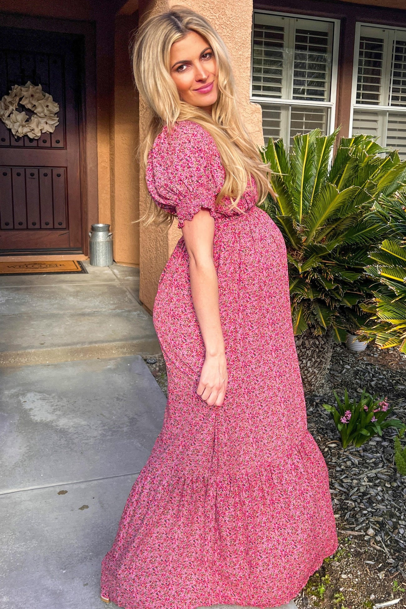 Pregnant woman with long blonde hair wearing a pink floral maternity maxi dress standing outside a house