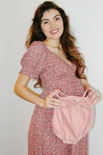 woman wearing a pink floral maternity dress and holding matching pink maternity bloomers, baby bump visible