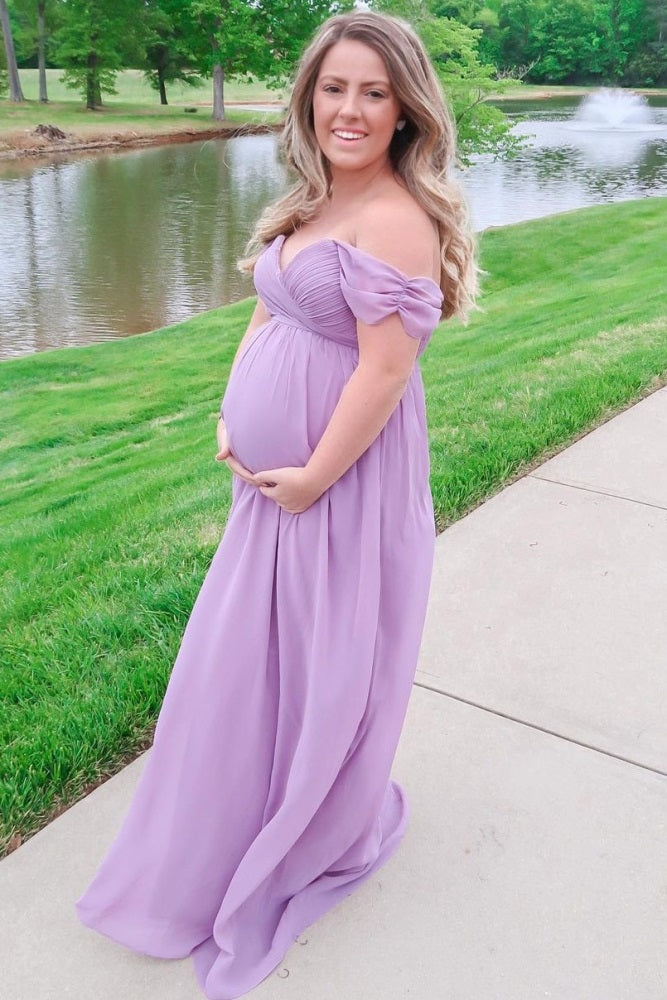 woman wearing a lavender off-shoulder maternity gown with flowing fabric, holding her baby bump by a serene pond