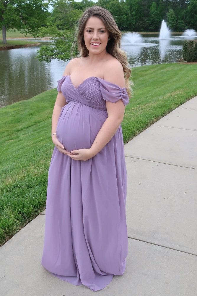 woman wearing a lavender off-the-shoulder maternity gown and holding her baby bump by a pond