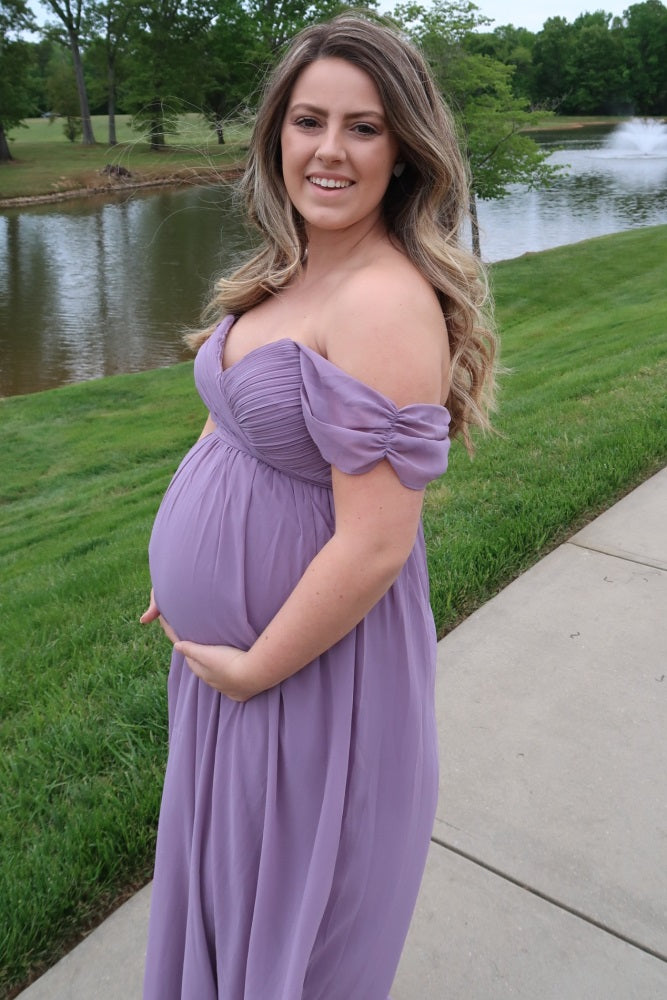 woman wearing a light purple off-the-shoulder maternity gown and holding her baby bump by a lakeside