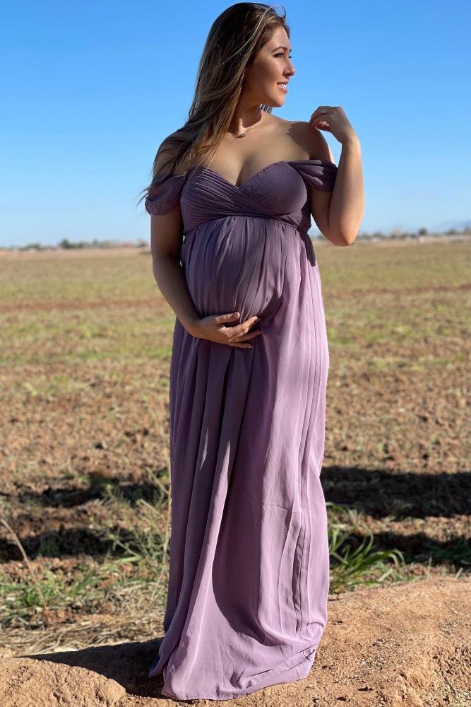 woman wearing a lavender off-the-shoulder maternity gown and holding her baby bump in a scenic outdoor setting
