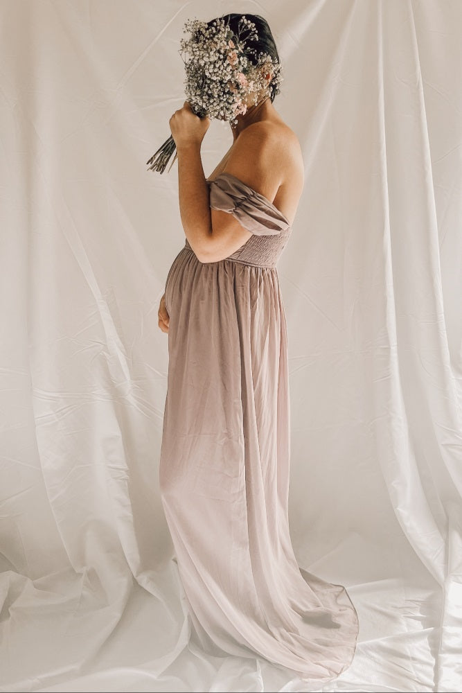 woman wearing a taupe off-the-shoulder maternity gown, holding a bouquet of flowers and revealing her baby bump