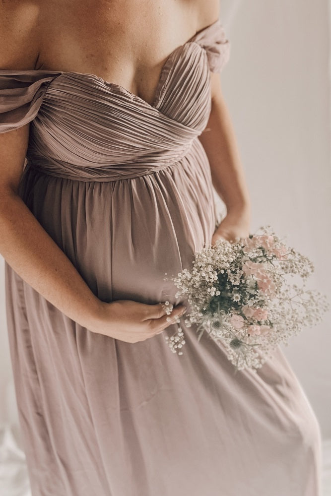 woman wearing a mauve off-the-shoulder maternity dress and holding her baby bump with a bouquet of flowers