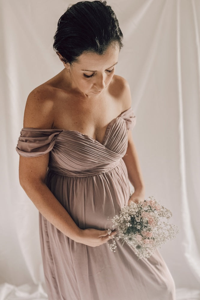 woman wearing a mauve off-the-shoulder maternity gown with pleated bodice, holding a baby bump and bouquet of flowers