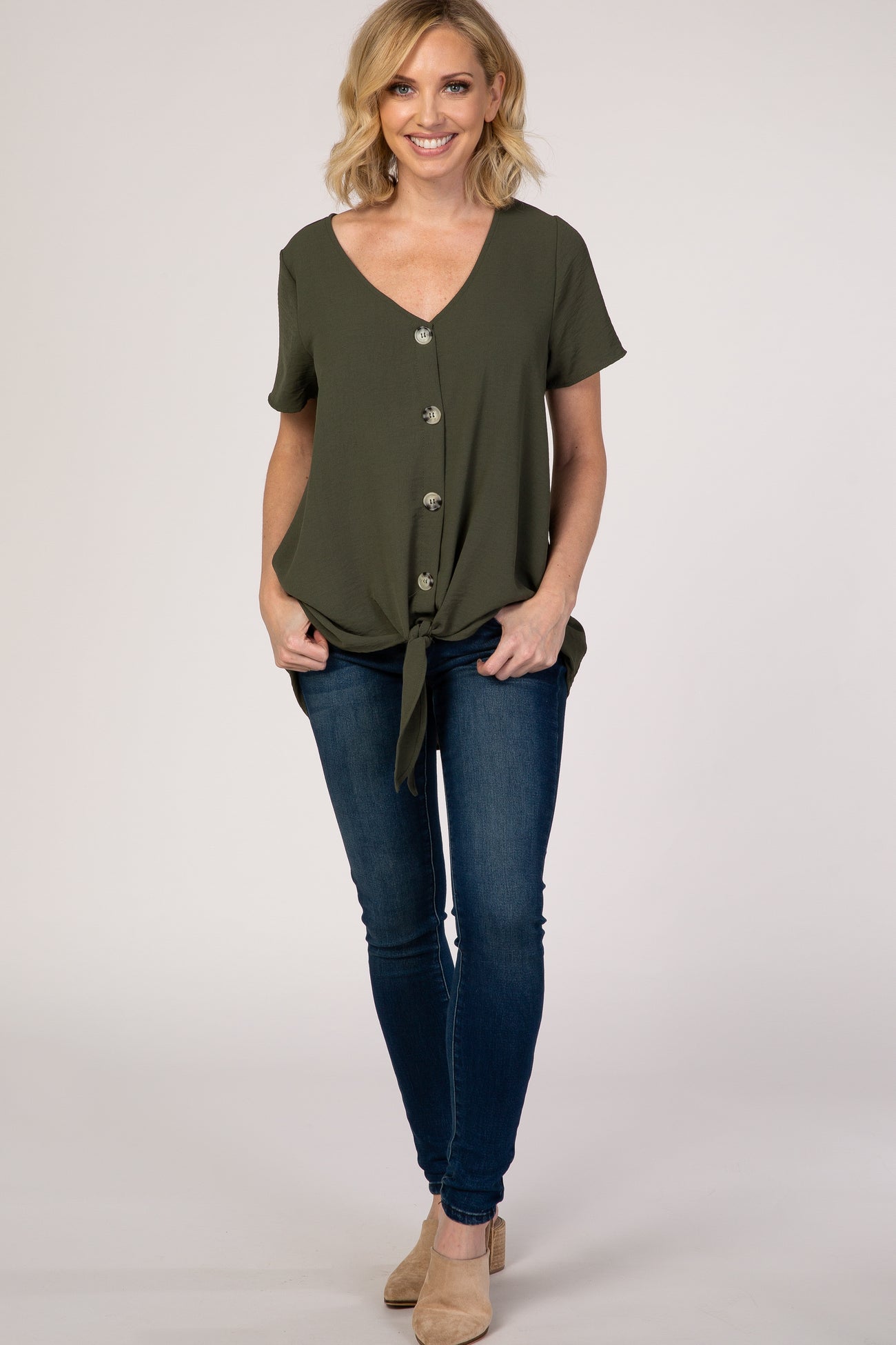 woman wearing an olive green short sleeve button-up top with a knot detail, paired with blue skinny jeans and beige heels