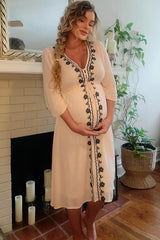 pregnant woman in cream embroidered maternity dress holding her belly standing by a fireplace