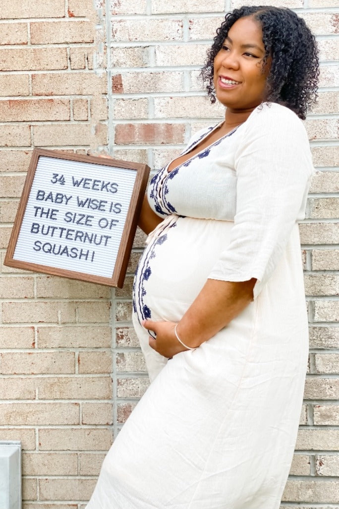 Pregnant woman in white dress holding a sign saying 34 weeks baby is the size of a butternut squash