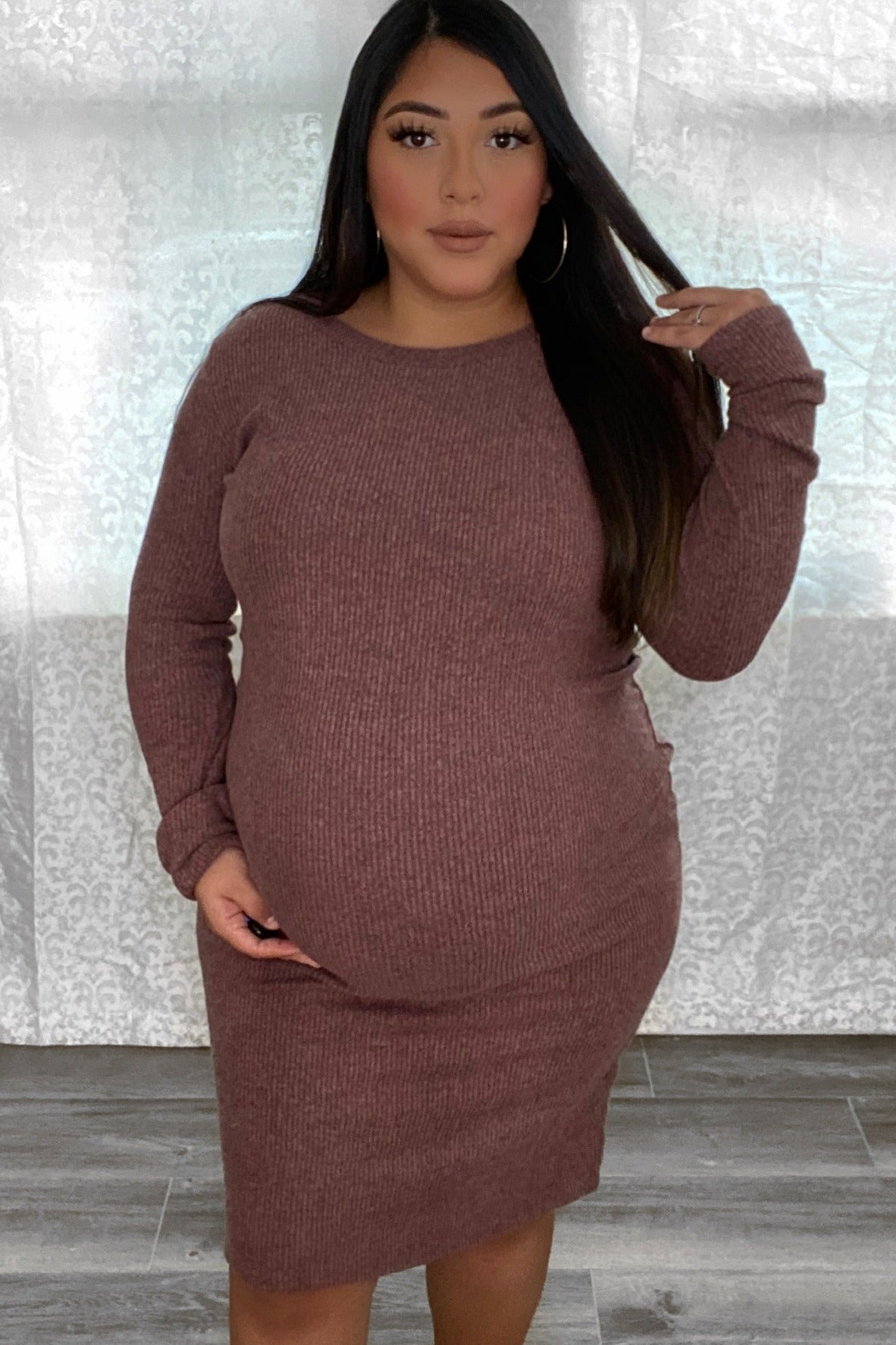 Pregnant woman wearing a long-sleeved mauve ribbed maternity dress standing in front of a white curtain