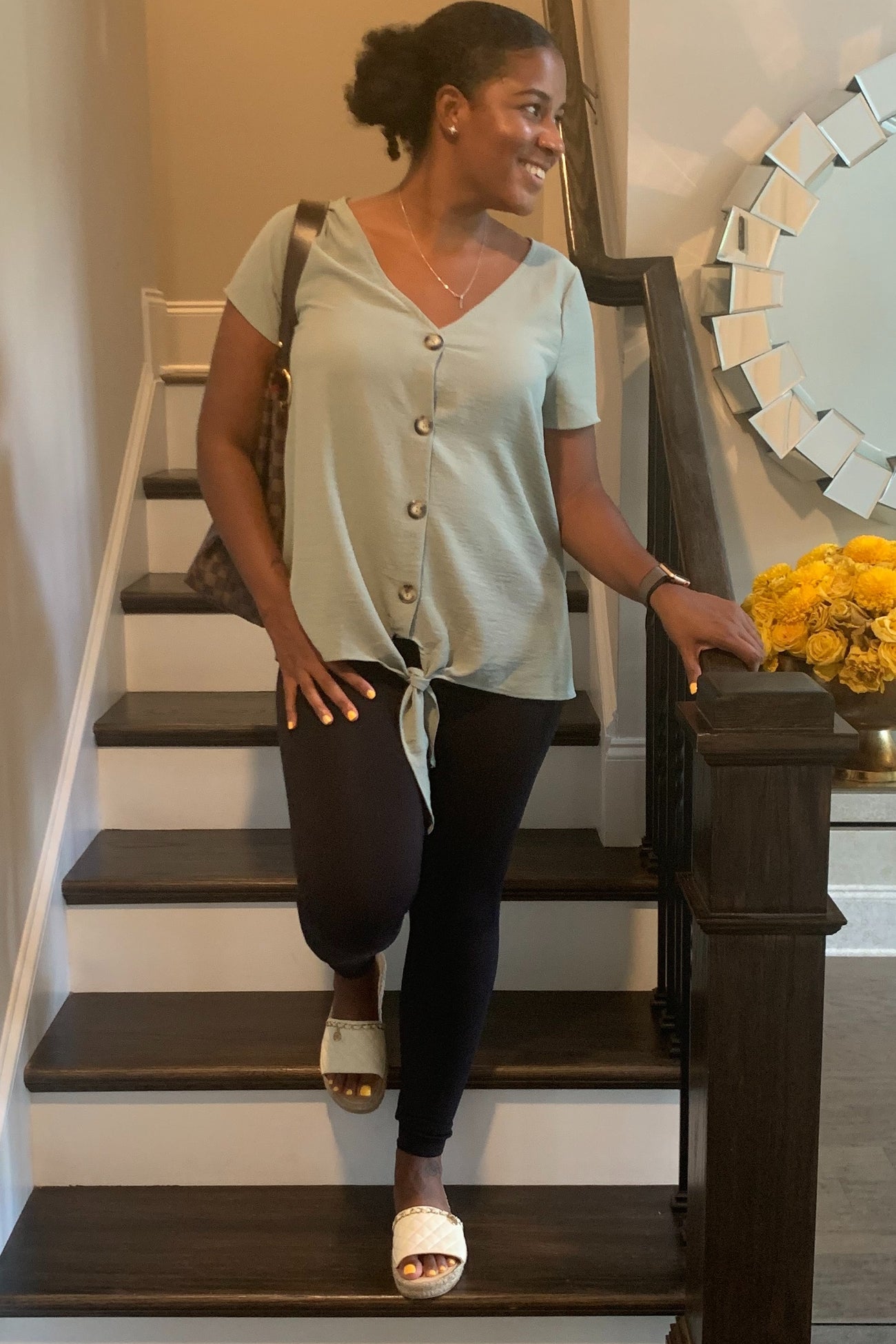 woman wearing a light green short sleeve top with buttons and a tie front, paired with black leggings and sandals, standing on stairs