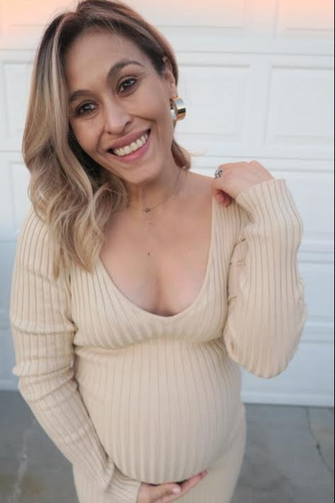 woman wearing a beige ribbed maternity dress and holding her baby bump, smiling at the camera