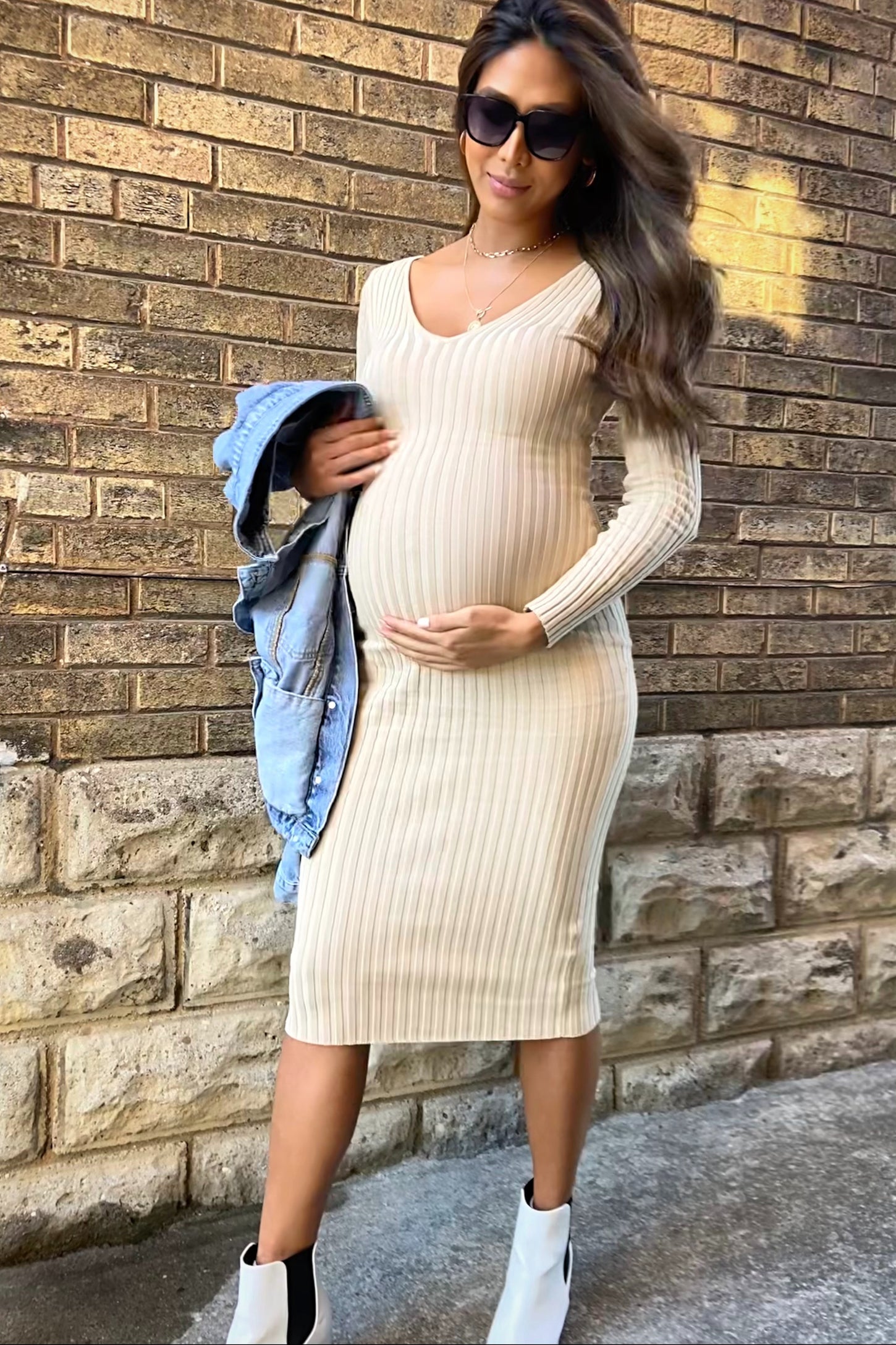 woman wearing a beige ribbed long sleeve maternity dress and holding her baby bump with a denim jacket draped over her arm