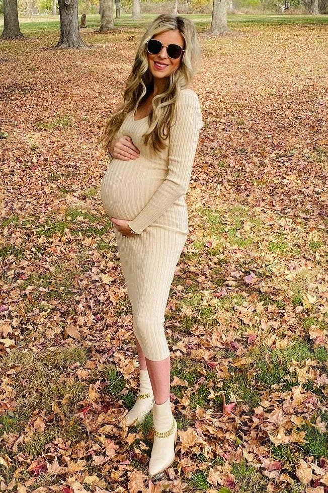 woman wearing a beige ribbed maternity dress and showing her baby bump, standing in a field of autumn leaves