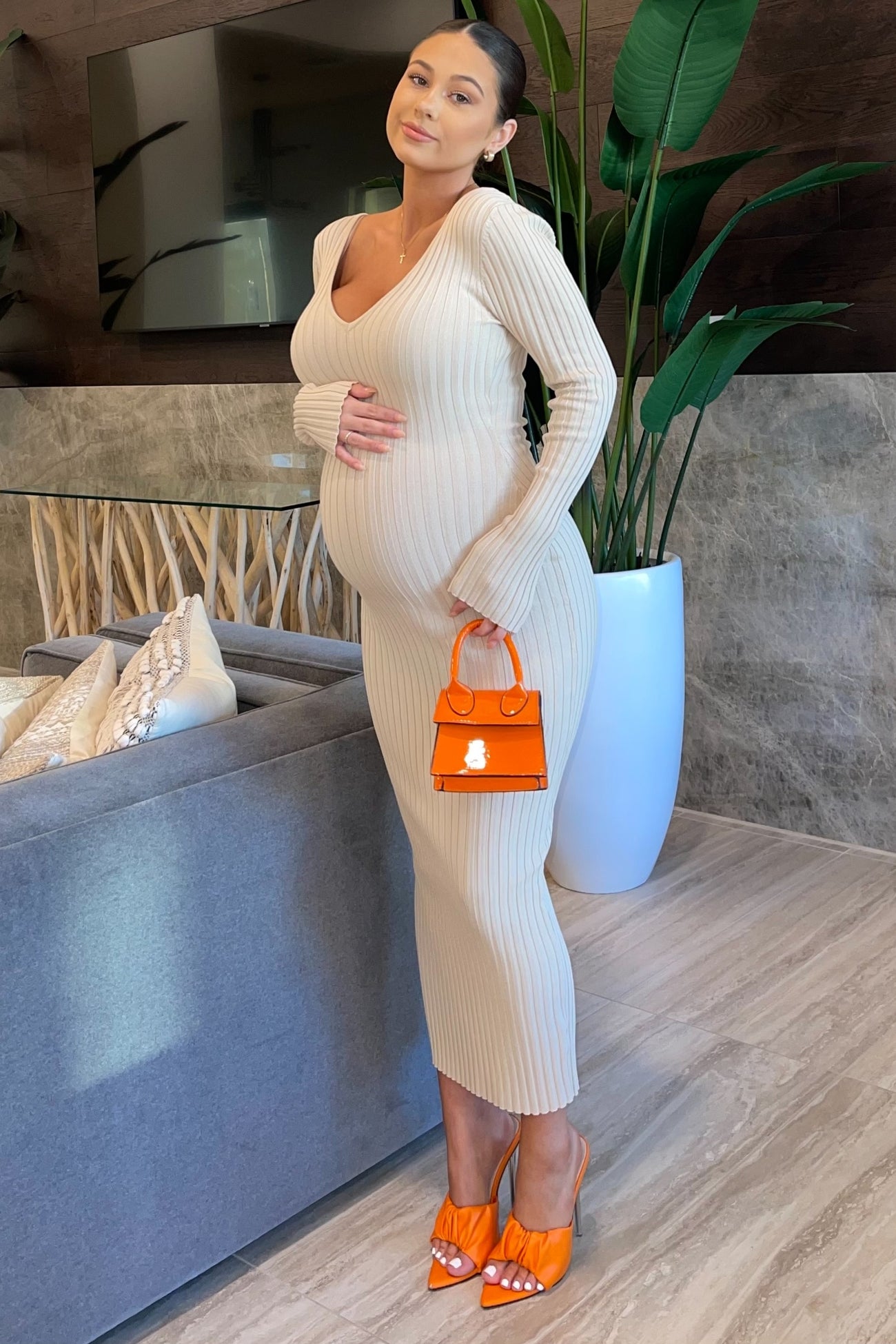woman wearing a beige ribbed maternity dress and holding an orange handbag, showing her baby bump