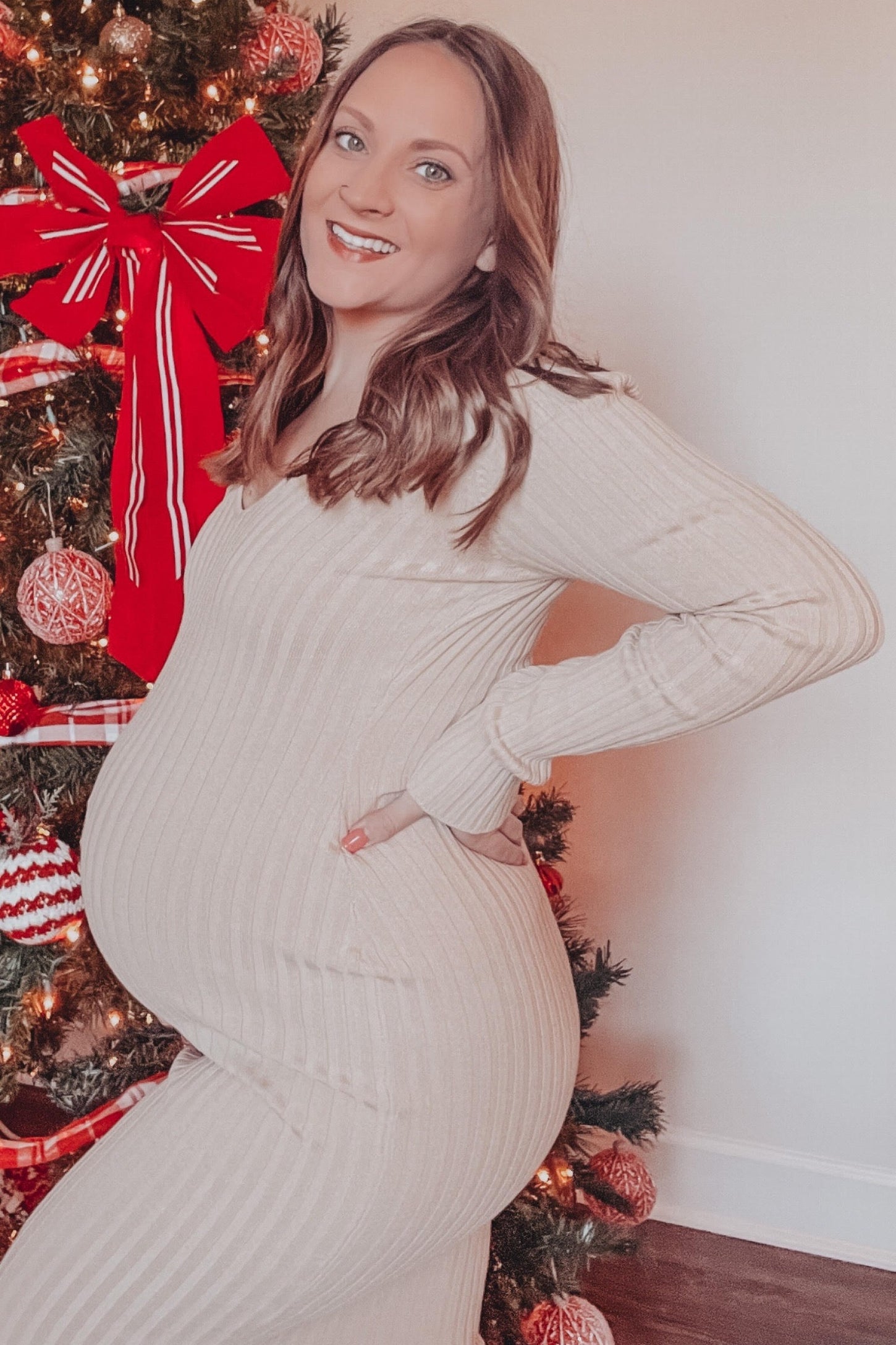 woman wearing a beige ribbed maternity dress and showing her baby bump in front of a decorated Christmas tree