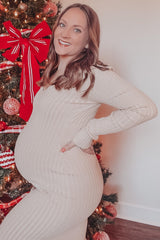 woman wearing a beige ribbed maternity dress and showing her baby bump in front of a decorated Christmas tree